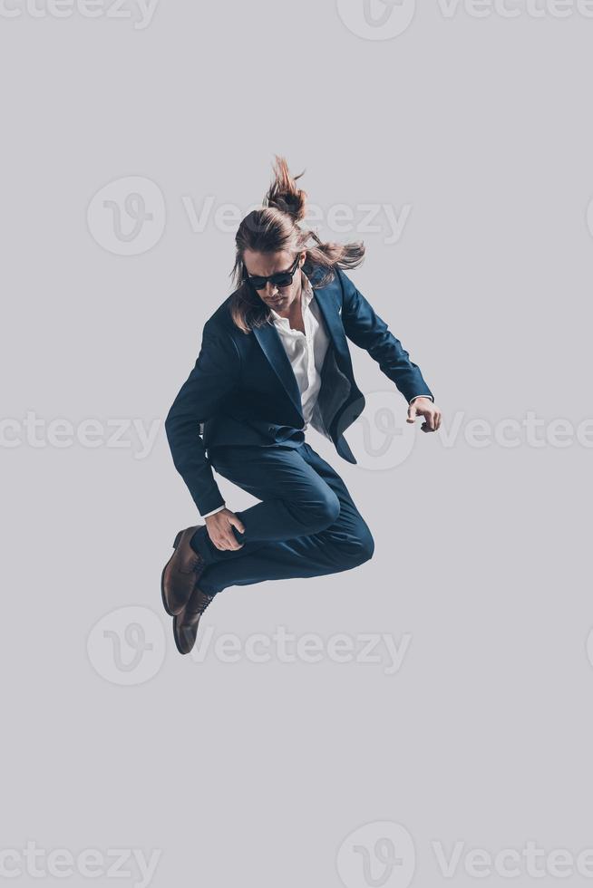 Mid-air style. Handsome young man in full suit and sunglasses jumping against grey background photo