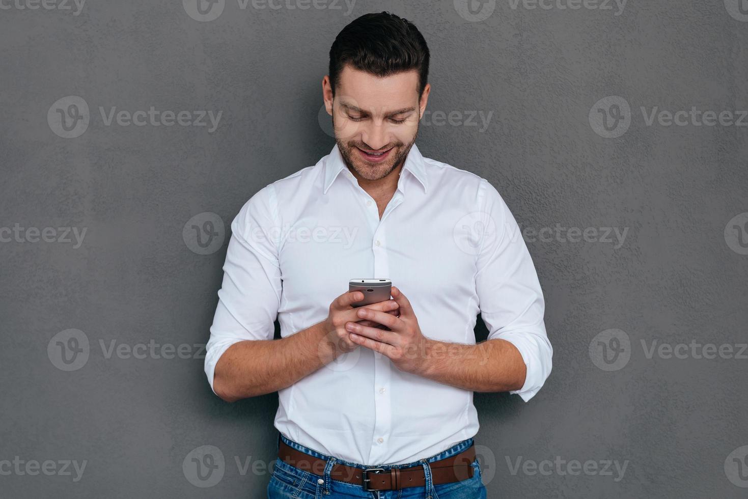 Texting business message. Handsome young man talking on the holding smart phone and looking at it with smile while standing against grey background photo