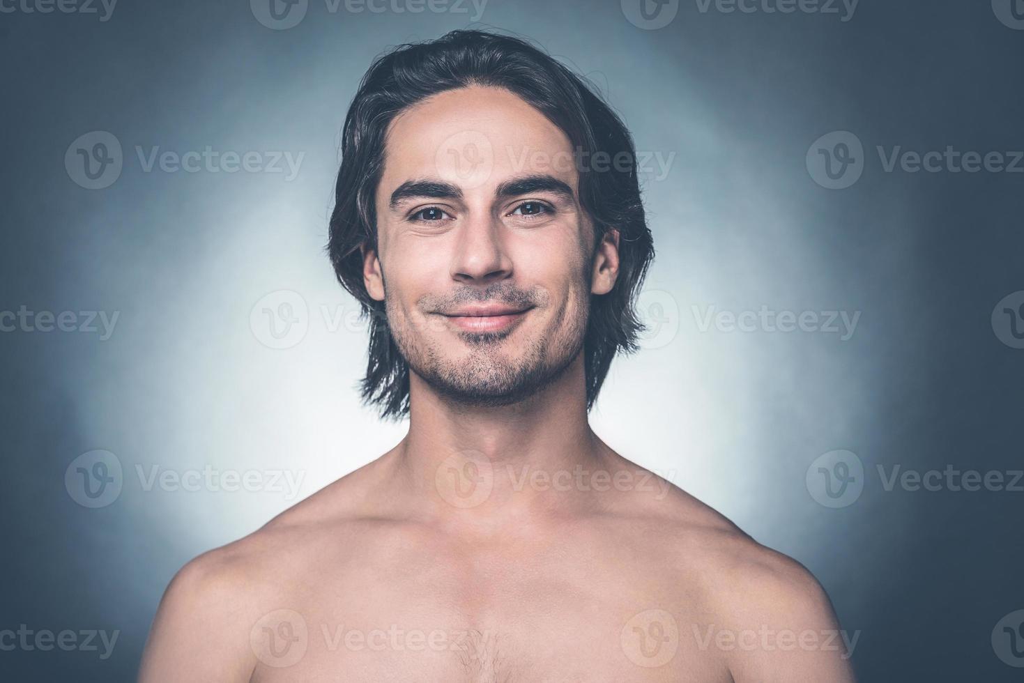 Confident and handsome. Portrait of young shirtless man looking at camera and smiling while standing against grey background photo