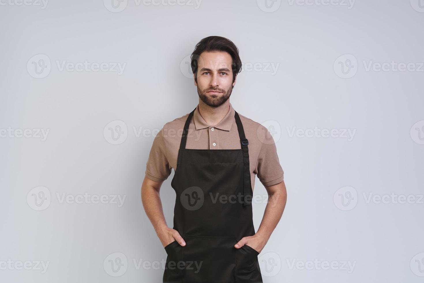 Handsome young man in apron looking at camera and holding hands on hip while standing against gray background photo