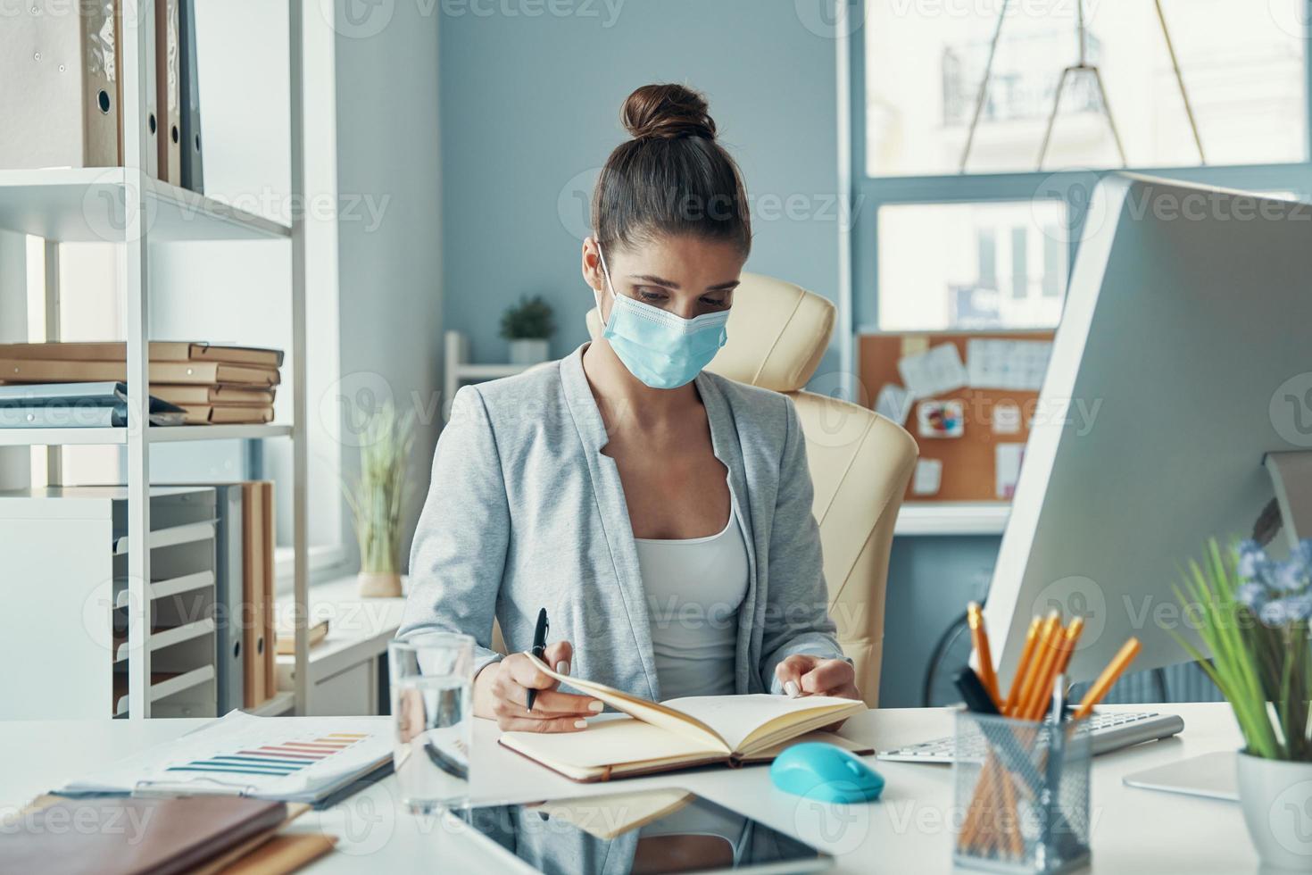Beautiful young woman in smart casual wear writing something down while sitting in the office photo