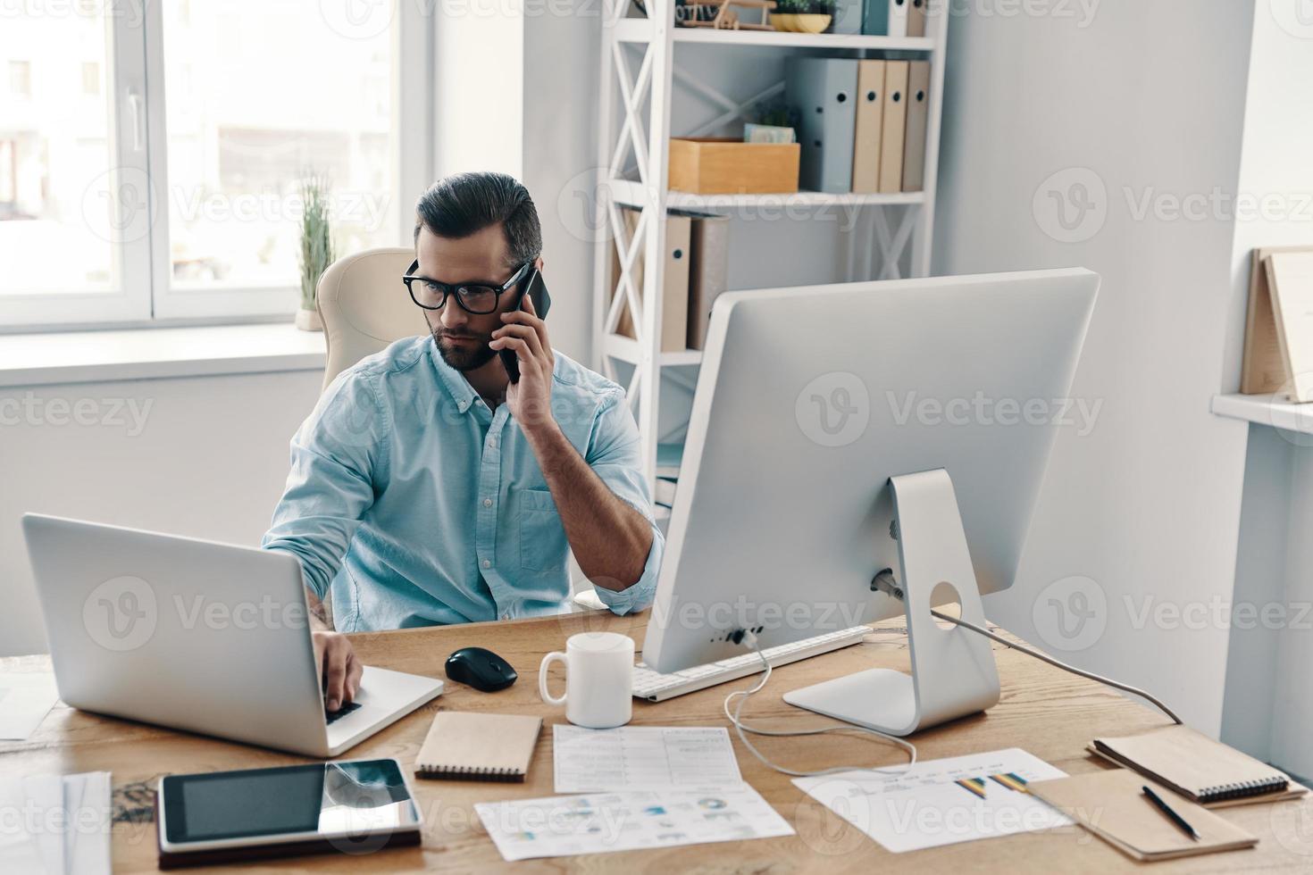 Working hard. Top view of young modern businessman talking on the phone and working using computer while sitting in the office photo