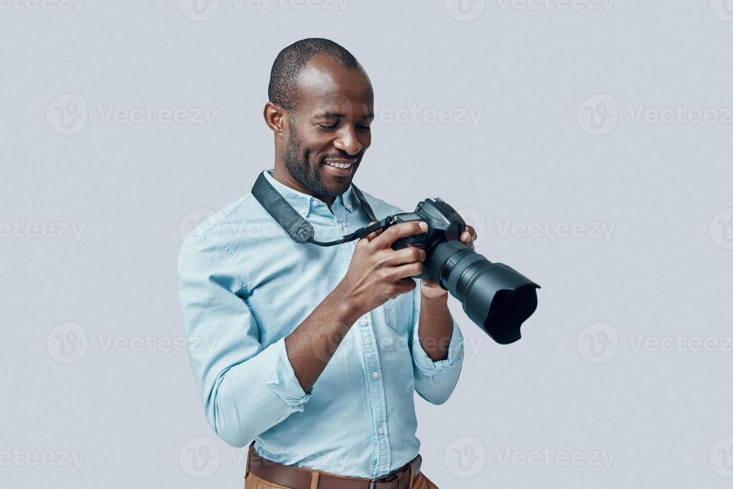Happy young African man using digital camera and smiling while standing against grey background photo