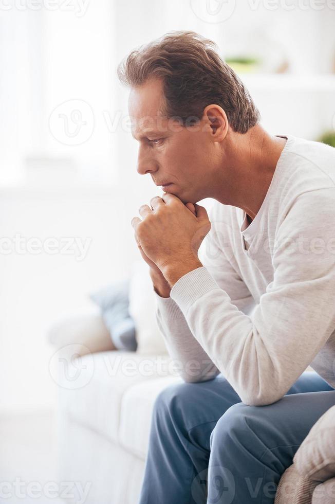 Feeling worried about... Side view of mature man holding hands on chin and looking worried while sitting on couch at home photo