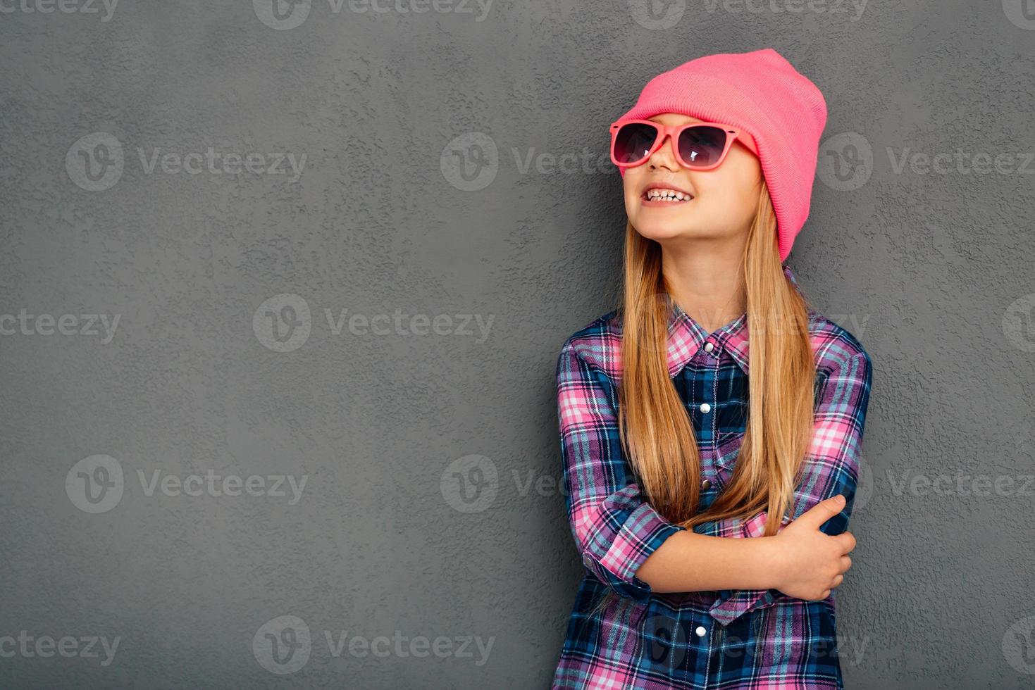 elegante y lindo. niña alegre con gafas de sol con los brazos cruzados y mirando hacia arriba con una sonrisa mientras se enfrenta a un fondo gris foto