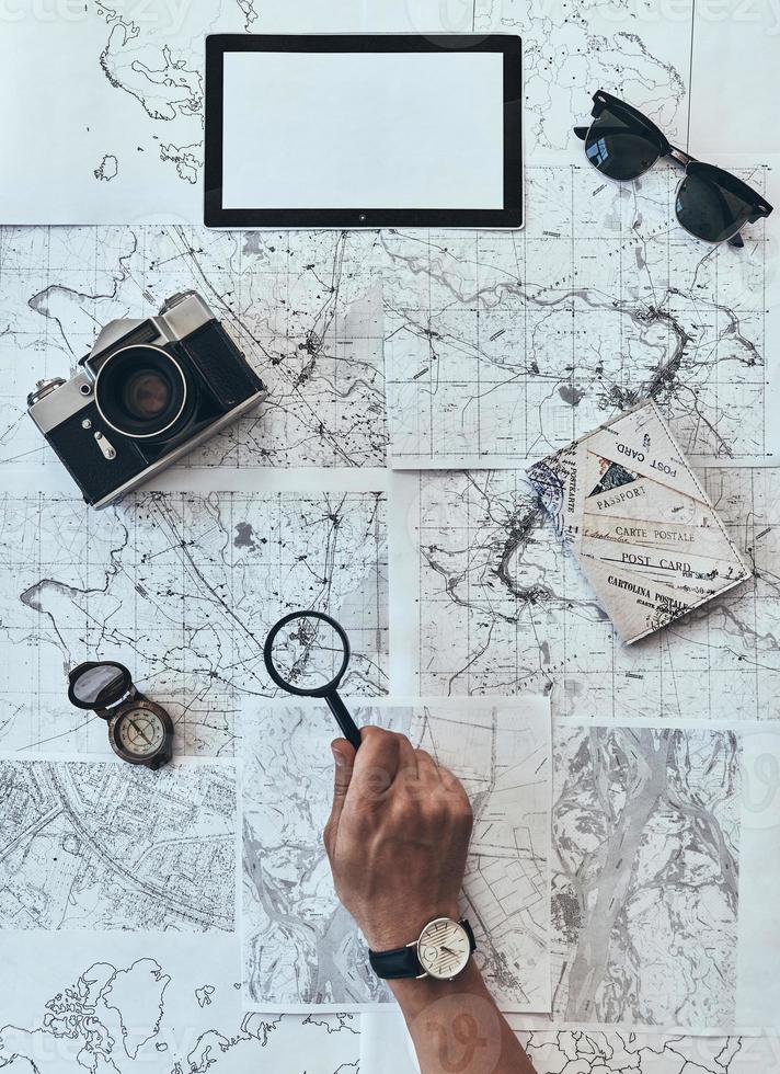 Trying to see every detail. Close up top view of man using magnifying glass on map with sunglasses, photo camera, compass, passport lying around