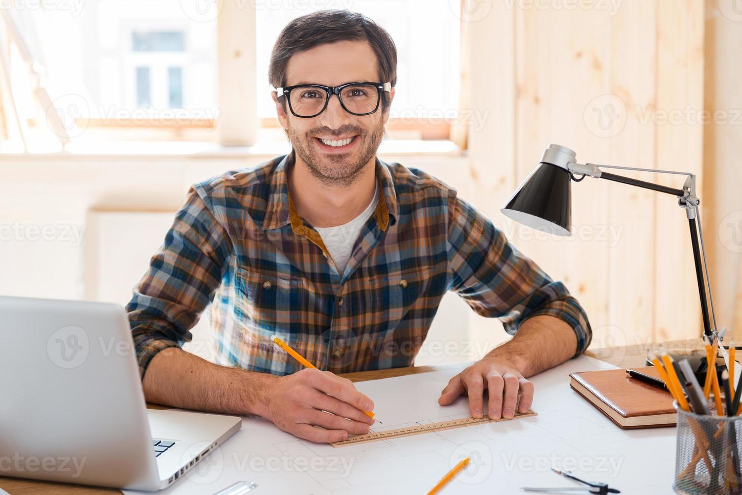 My space to create. Handsome young man designing project and smiling at camera while sitting at his working place photo