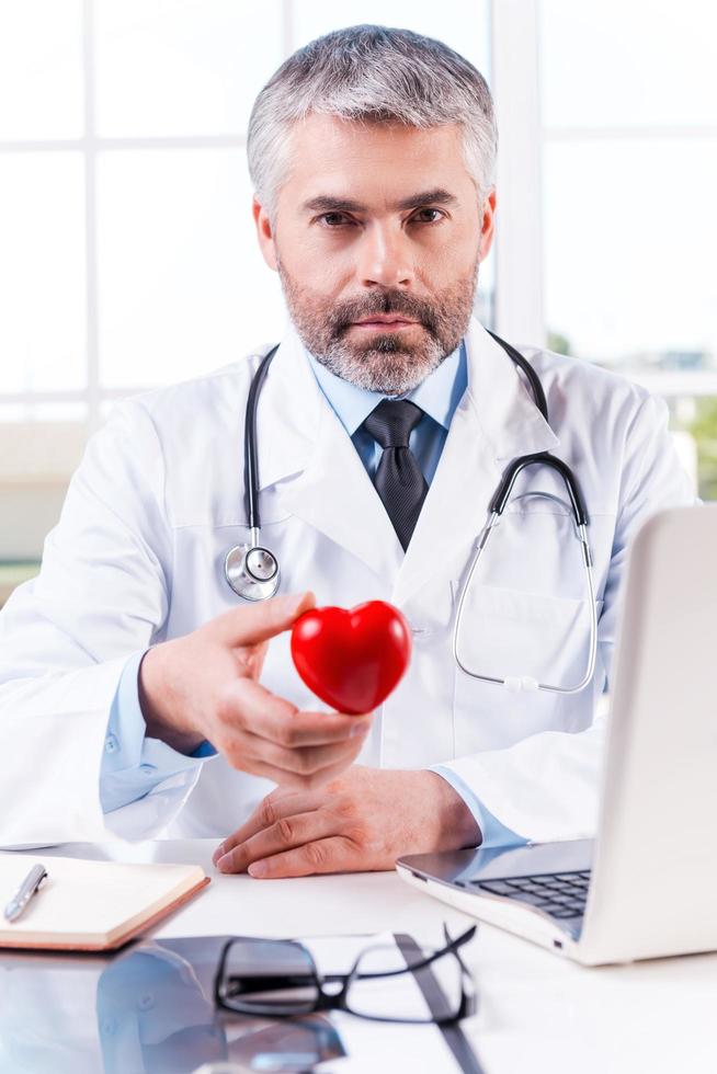 Taking care of your heart. Mature grey hair doctor holding heart shape toy and looking at camera while sitting at his working place photo