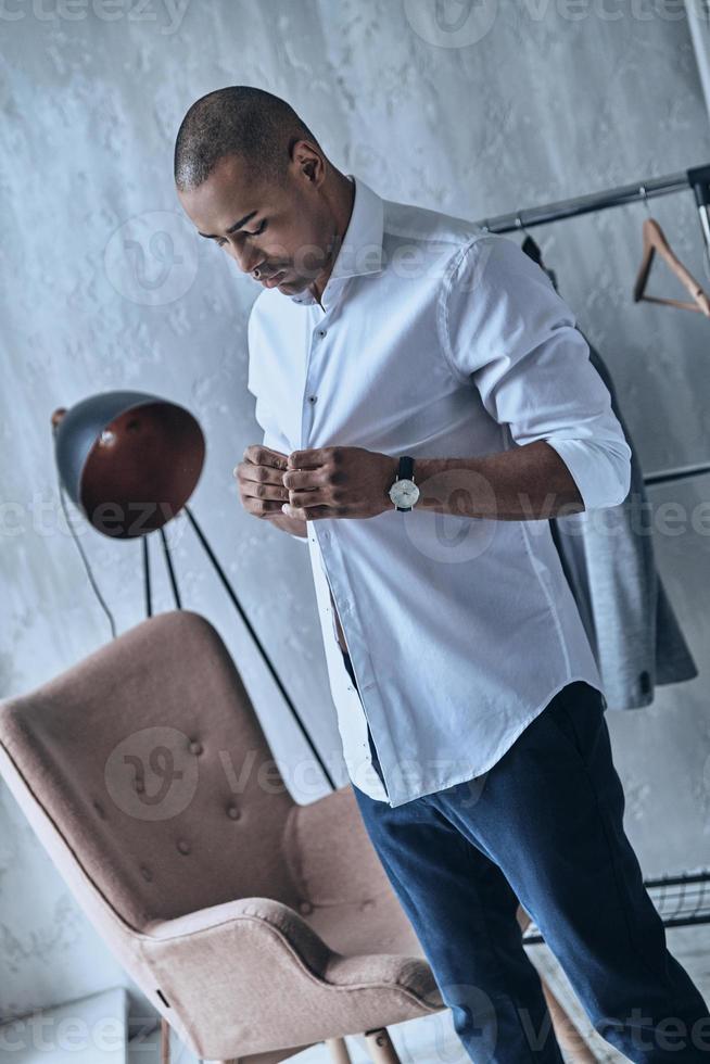 Confident in his style. Handsome young African man buttoning his shirt while standing at home photo