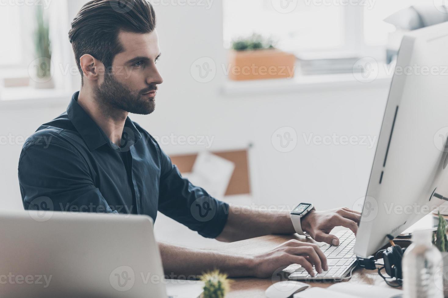 día de trabajo ocupado. un joven apuesto que trabaja en la computadora mientras está sentado en su lugar de trabajo en la oficina de su casa foto