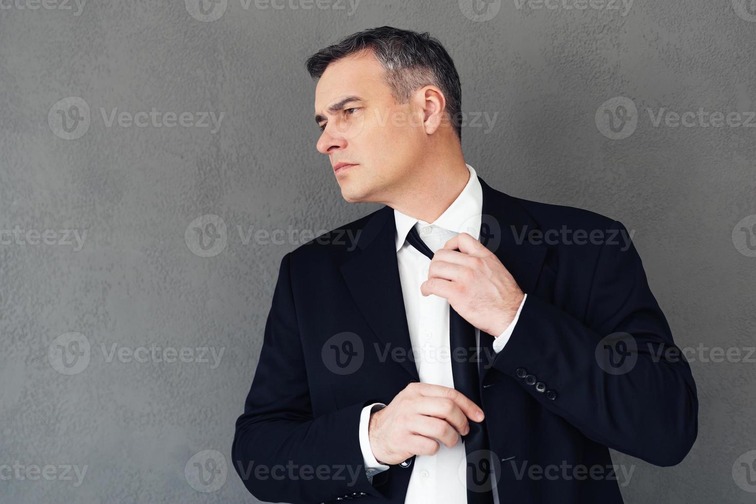 Tired from business. Mature businessman adjusting his tie while standing against grey background photo