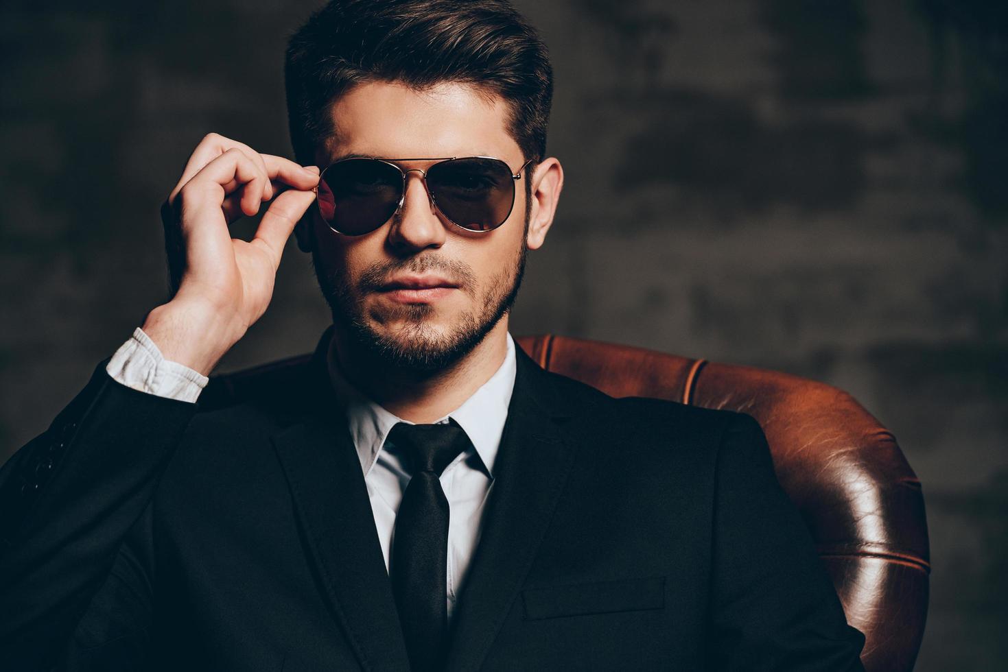 Portrait of confidence.Portrait of young handsome man in suit adjusting his sunglasses and looking at camera while sitting in leather chair against dark grey background photo