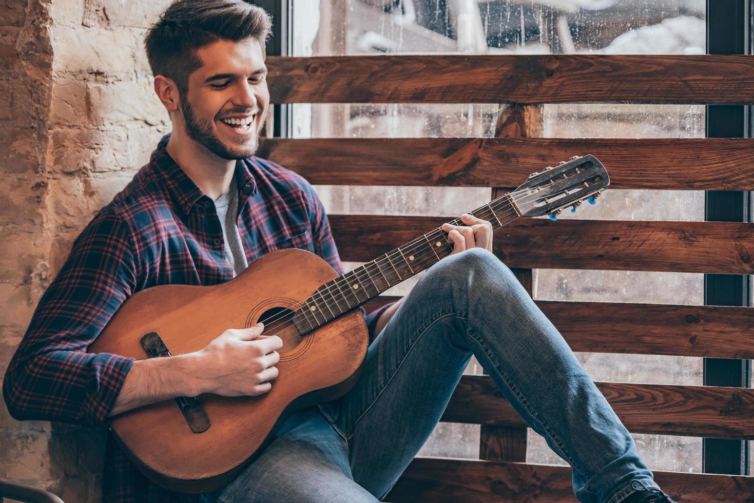 guitarrista alegre. un joven apuesto y alegre tocando la guitarra y sonriendo mientras se sienta en el alféizar de la ventana foto