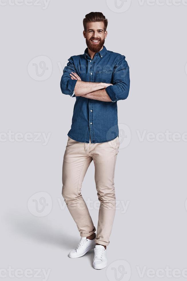 Confident in his style. Full length of handsome young man looking at camera and keeping arms crossed while standing against grey background photo