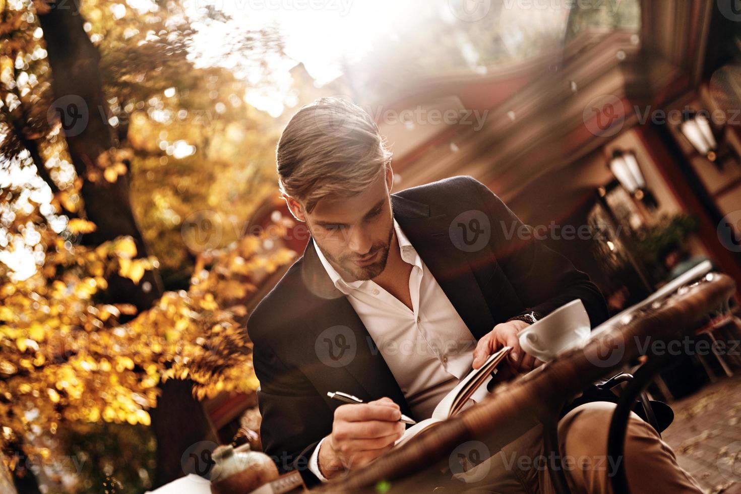 Everything must be planned. Good looking young man in smart casual wear writing something down in his personal organizer while sitting in restaurant outdoors photo