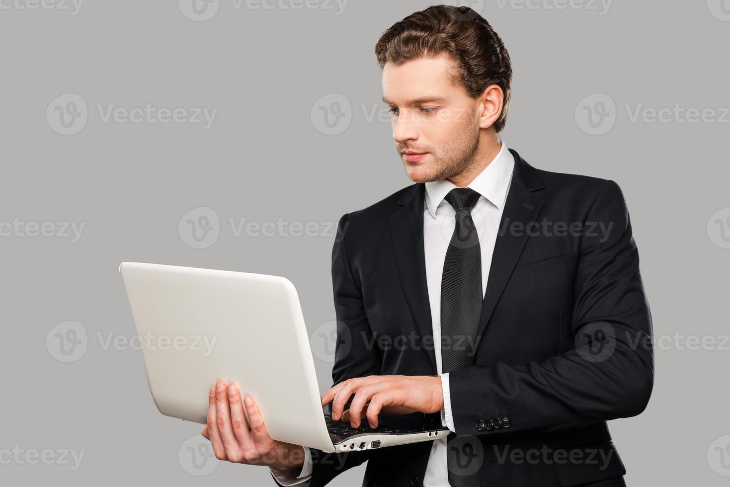 Businessman with laptop. Confident young man in formalwear working on laptop while standing against grey background photo