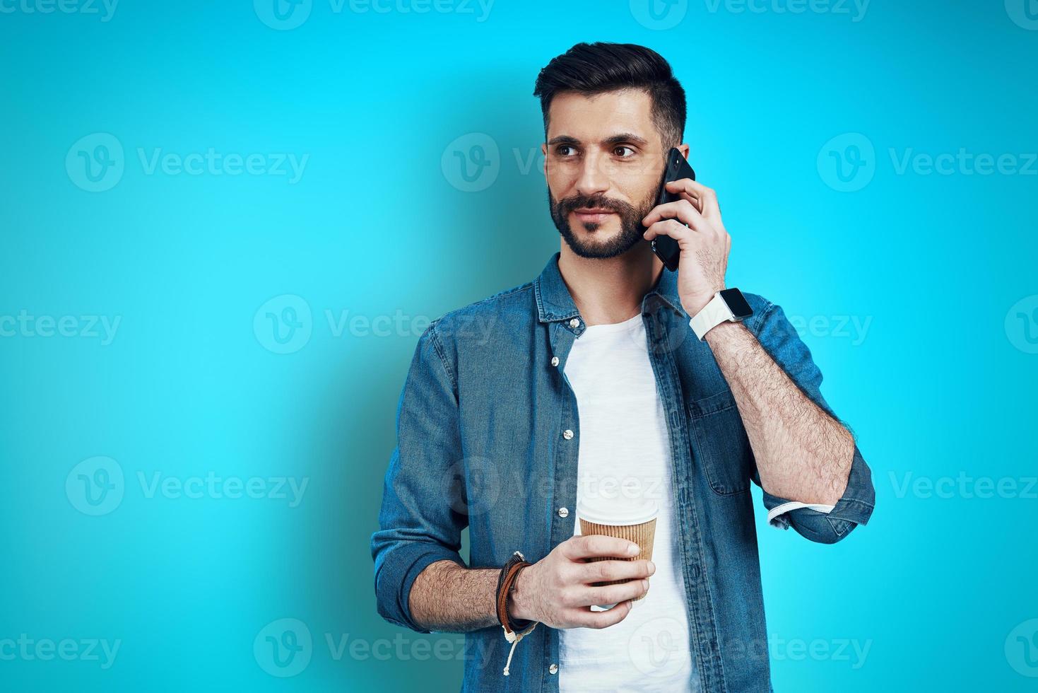 Good looking young man in casual wear talking on smartphone while standing against blue background photo