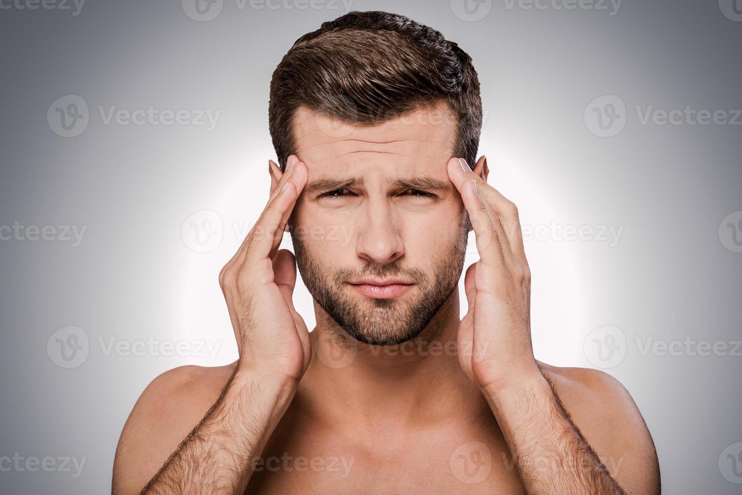Feeling headache. Portrait of frustrated young shirtless man looking at camera and touching head with hands while standing against grey background photo