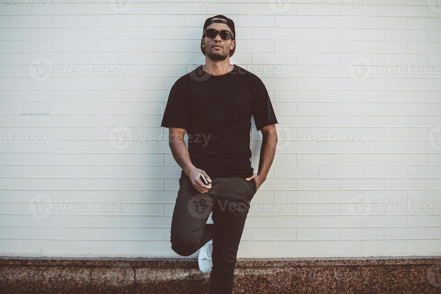 Cool and handsome. Handsome young African man in casual clothes leaning at the brick wall and holding smart phone photo