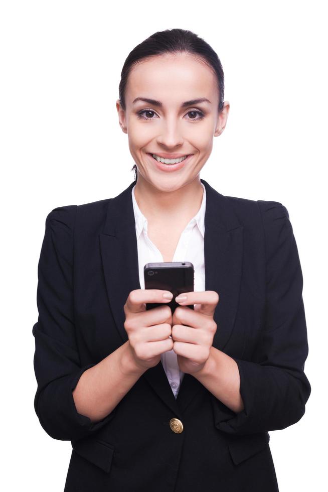 Business lady with mobile phone. Cheerful young woman in formalwear holding mobile phone and smiling while standing isolated on white photo