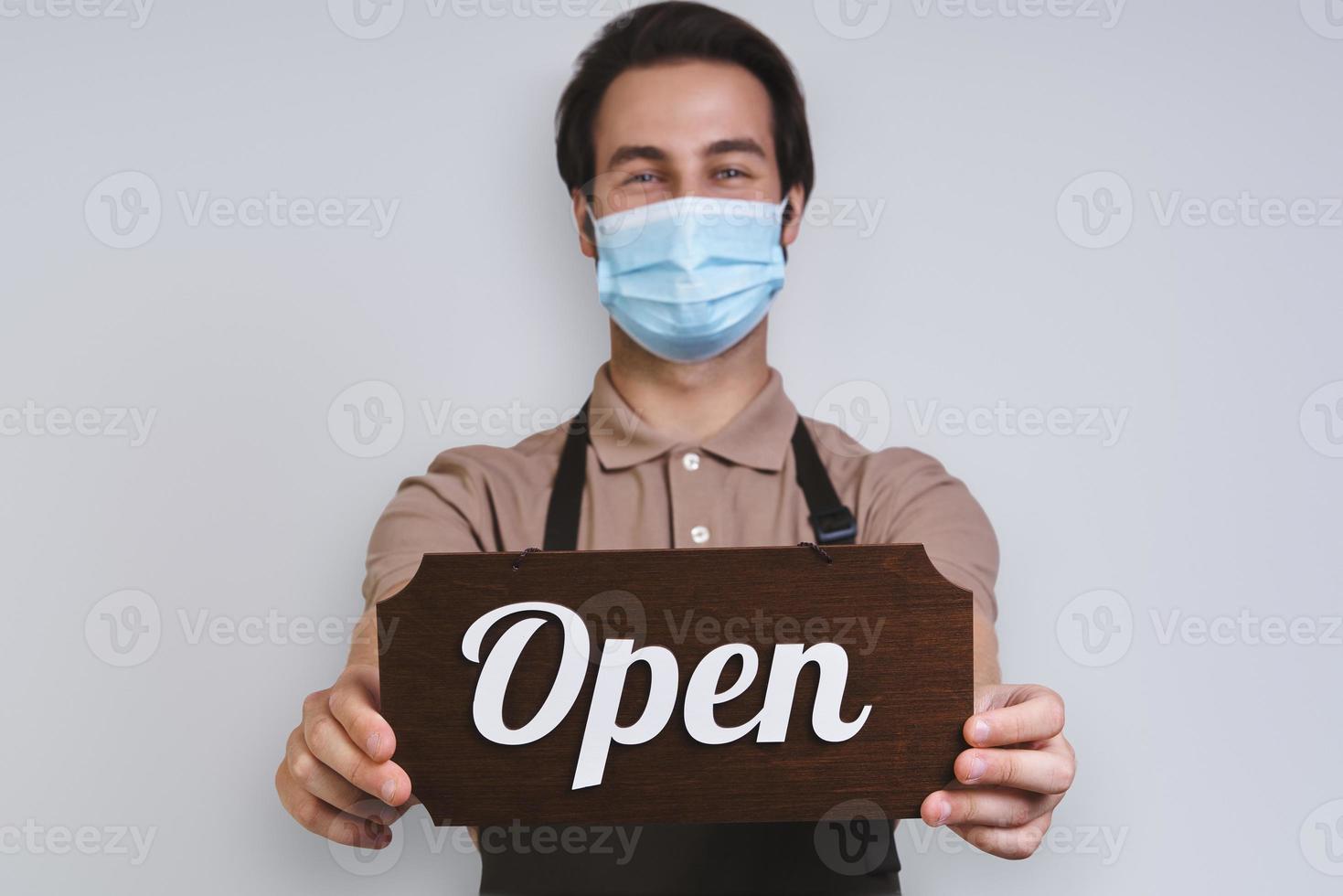 Handsome young man in apron wearing protective face mask and holding open sign while standing against gray background photo