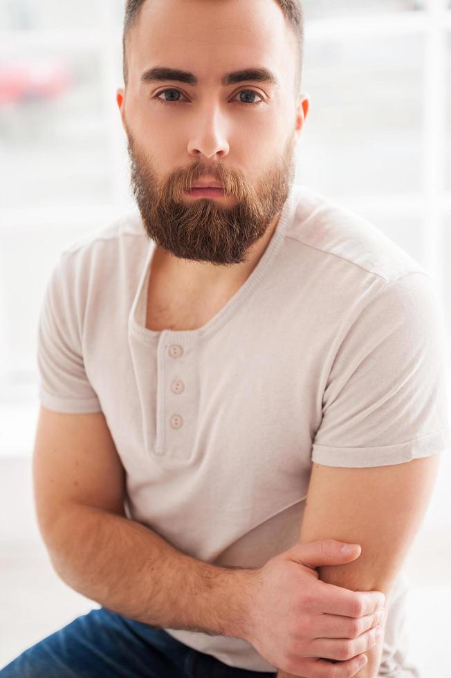 Beard handsome. Thoughtful young man in t-shirt and looking at camera while sitting on the chair photo