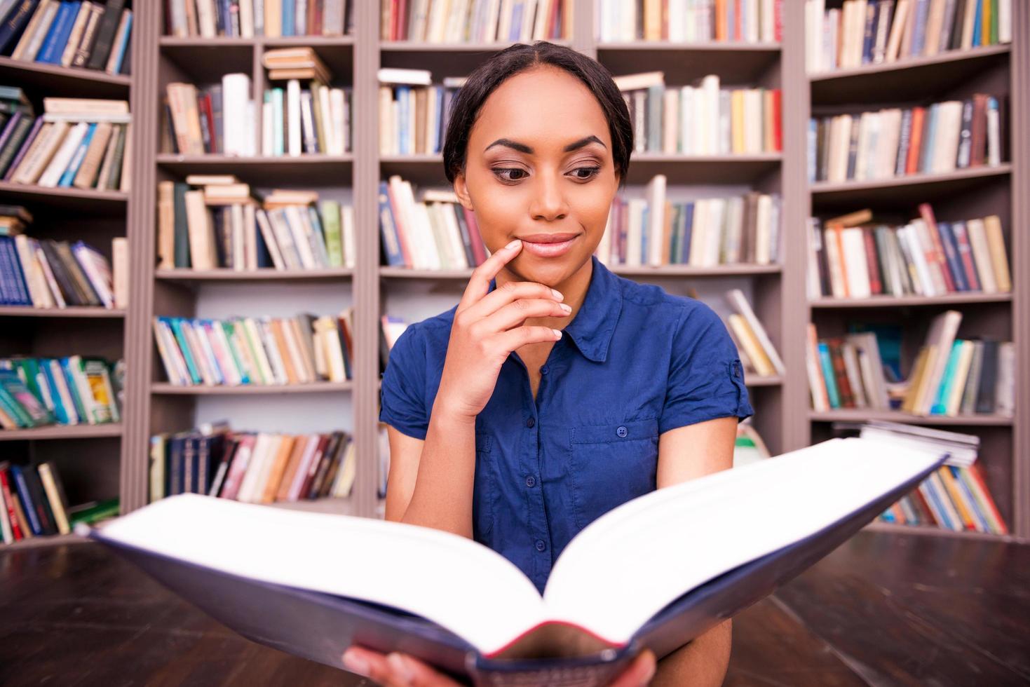 Beautiful bookworm. Thoughtful African female student reading book and holding hand on chin while sitting on the floor in library photo