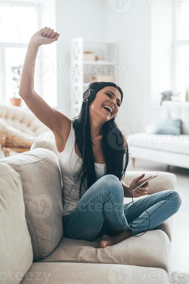 tiempo para la música hermosa mujer joven en auriculares sosteniendo un teléfono inteligente y gesticulando mientras se sienta en el sofá en casa foto