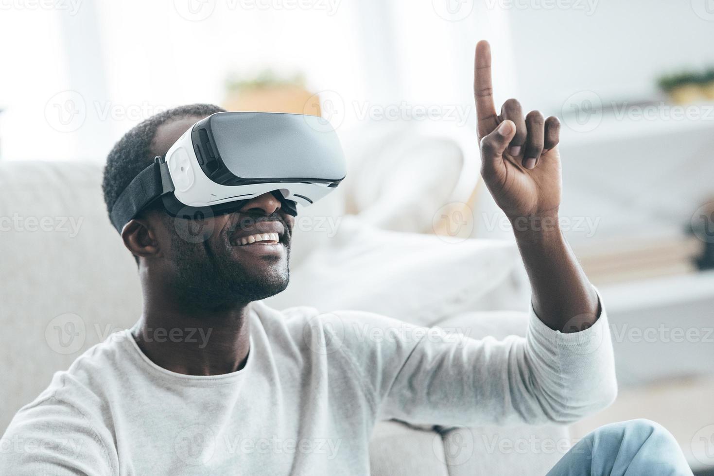 todo es tan real y apuesto joven africano con ropa elegante e informal apuntando a algún lado y sonriendo mientras se sienta en la alfombra en casa foto