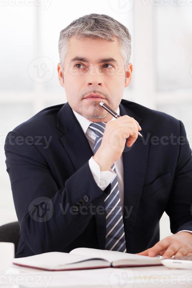 esperando inspiración. un anciano pensativo con ropa formal sosteniendo un bolígrafo en la barbilla y mirando hacia otro lado mientras estaba sentado en su lugar de trabajo foto