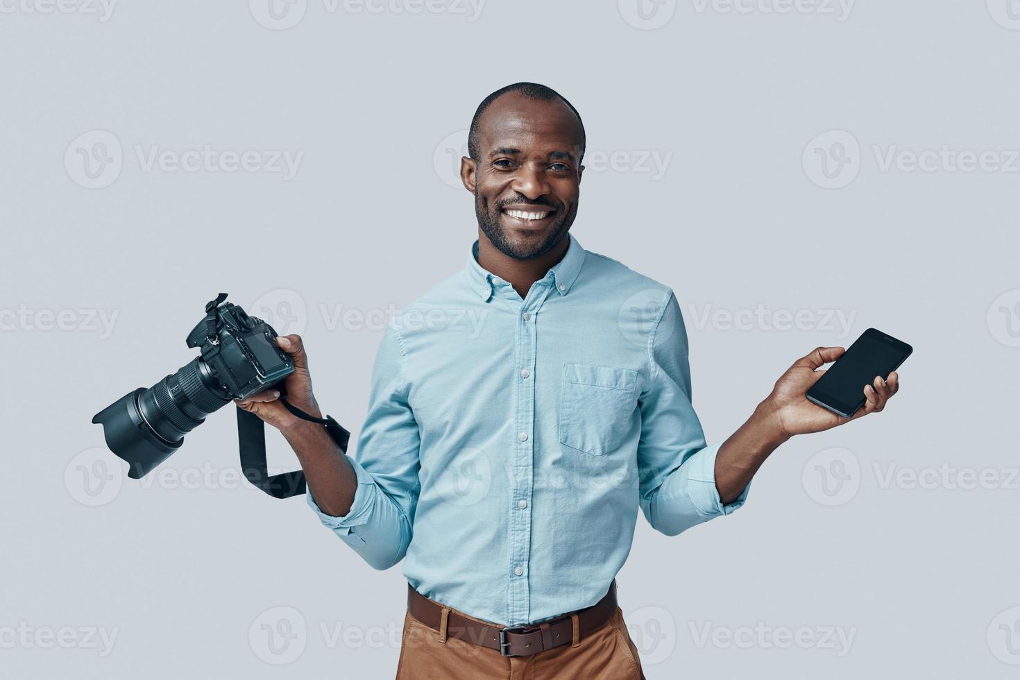 Happy young African man using digital camera and smiling while standing against grey background photo