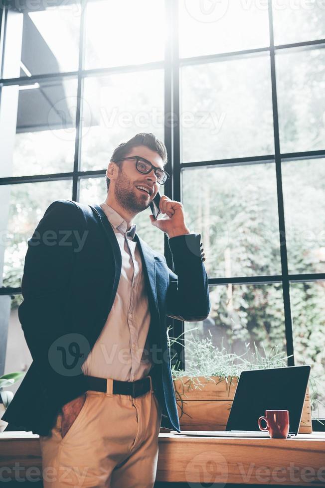 Good news from colleague. Low angle view of confident young man talking on the mobile phone and smiling while standing in front of the big window in office or cafe photo