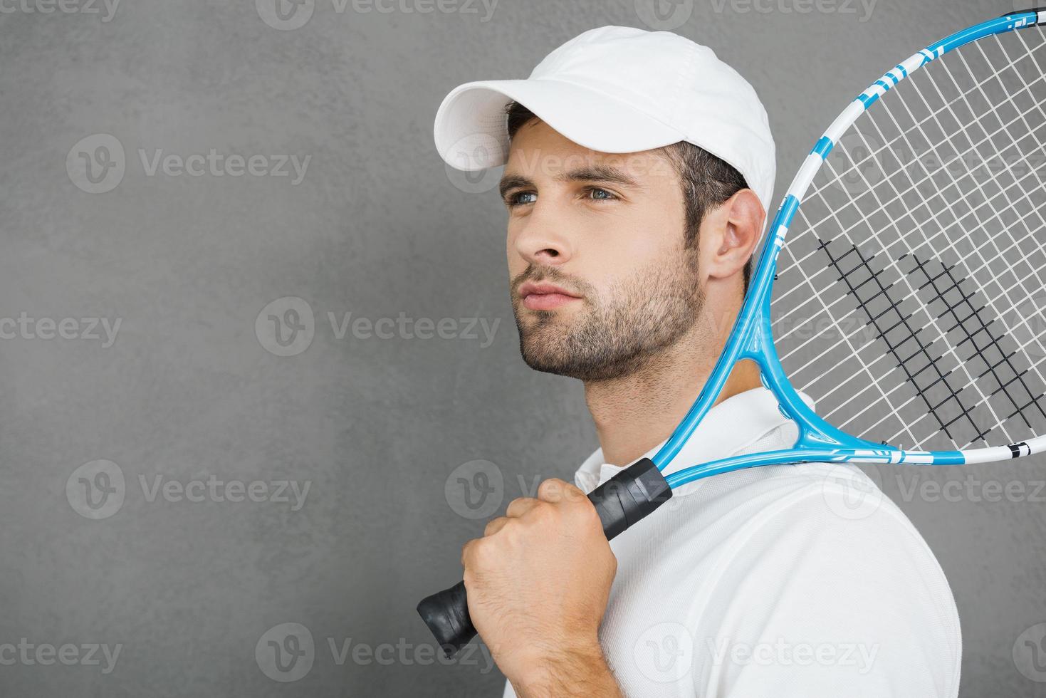 Tennis is his passion. Confident young man in sports clothes carrying tennis racket on his shoulder and looking away while standing against grey background photo