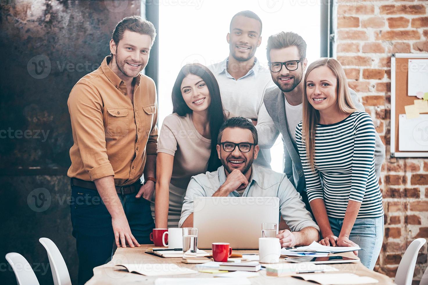 equipo creativo perfecto. grupo de seis jóvenes alegres mirando la cámara con una sonrisa mientras se inclinan a la mesa en la oficina foto