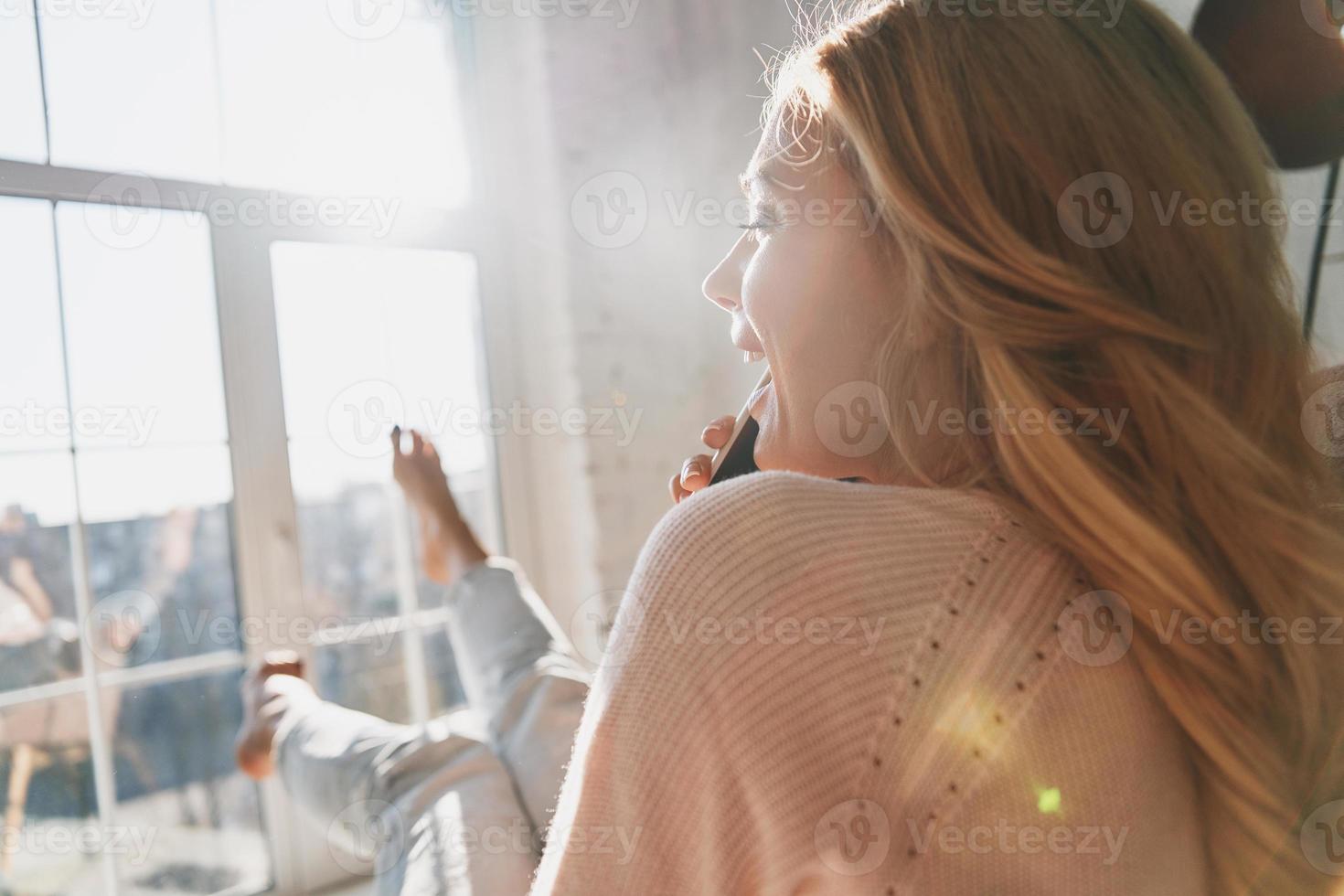 Rear view of beautiful young woman talking on her smart phone and smiling while sitting near the window at home photo