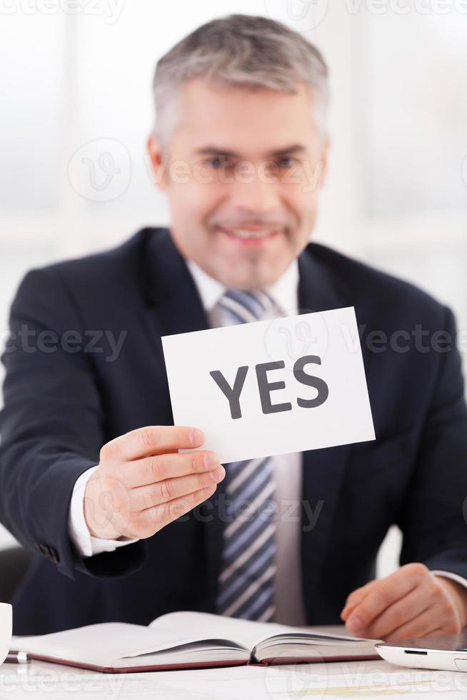 Yes Cheerful senior man in formalwear holding a paper with yes sign on it while sitting at her working place photo