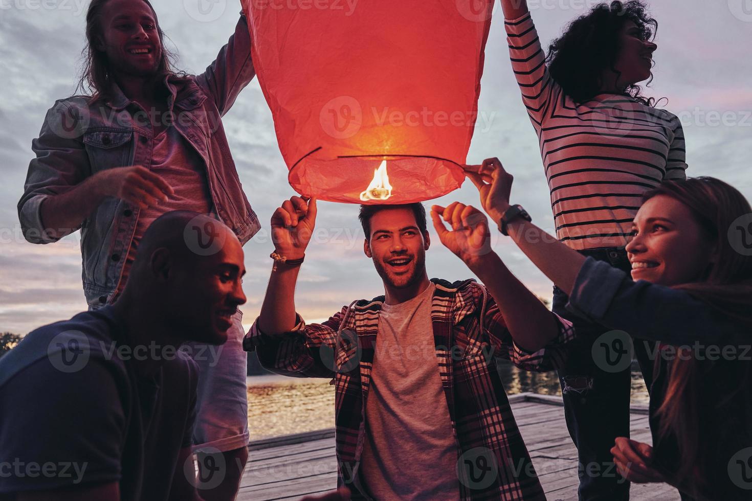 Young and free. Happy young people in casual wear lighting up a sky lantern while standing on the pier photo