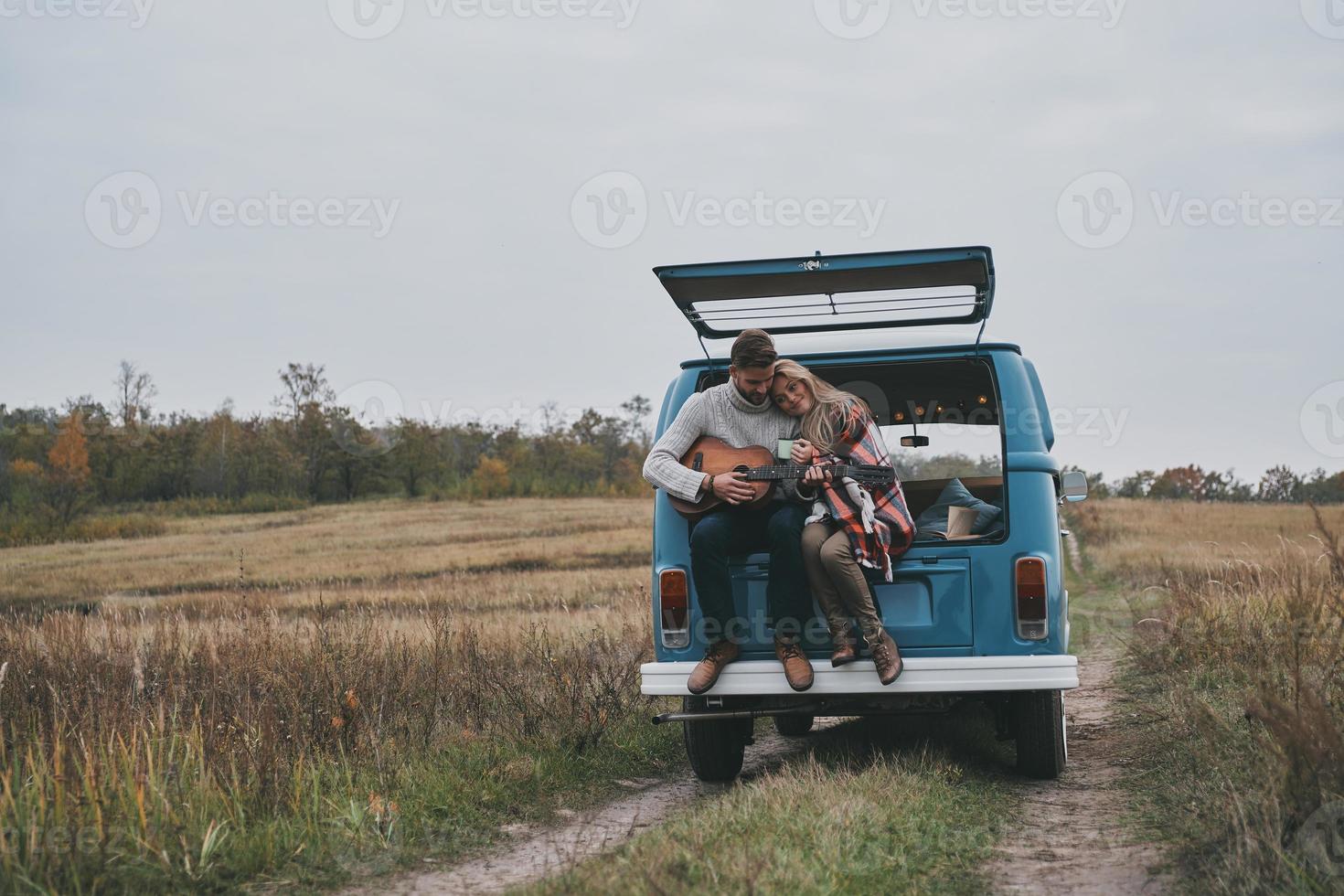 She is his muse.  Handsome young man playing guitar for his beautiful girlfriend while sitting in the trunk of blue retro style mini van photo