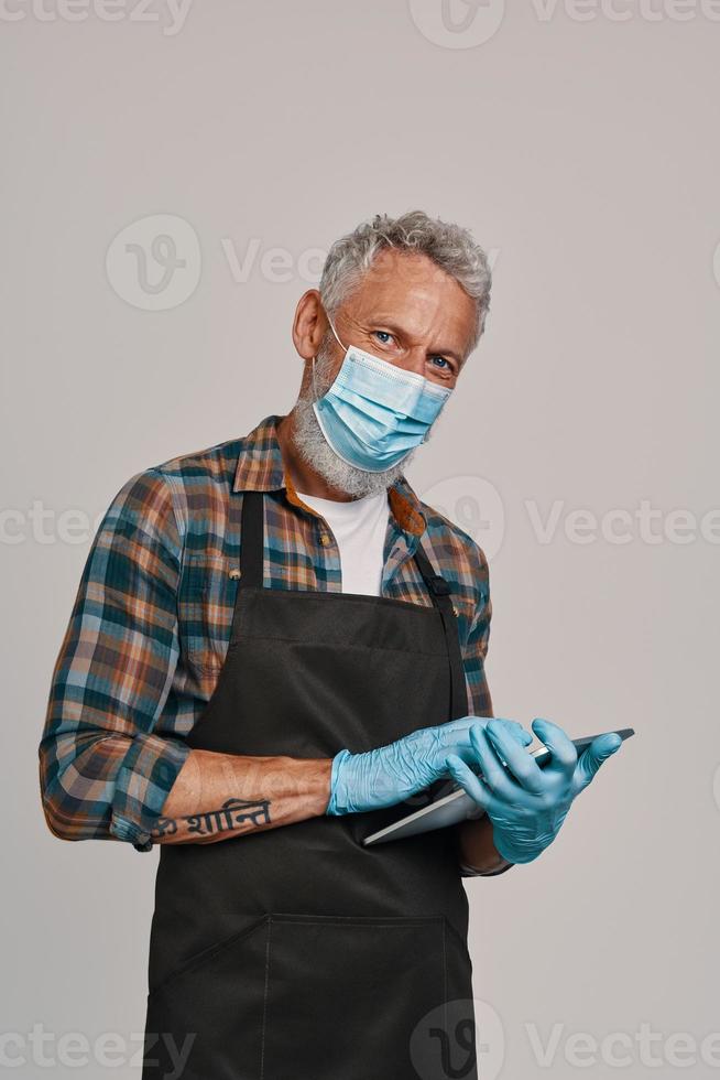 Senior man in apron and protective face mask using digital tablet photo