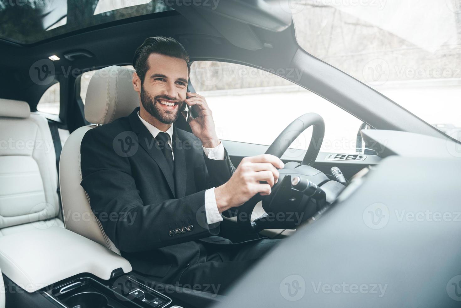 agradable charla de negocios. apuesto joven hombre de negocios hablando por su teléfono inteligente y sonriendo mientras está sentado en el asiento delantero foto