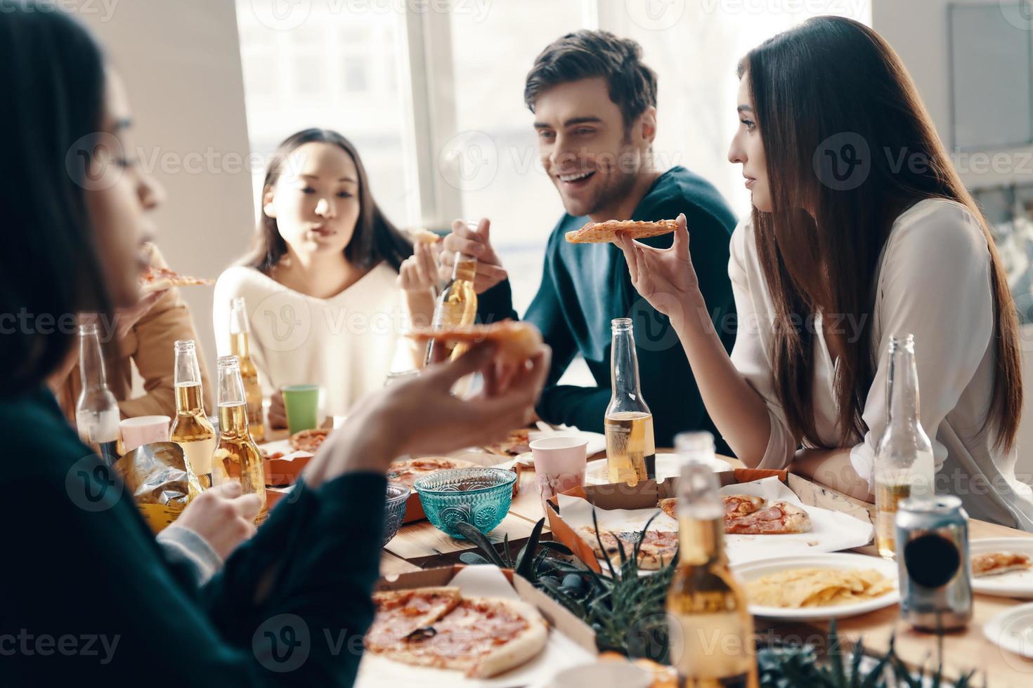 Great food and great people. Group of young people in casual wear eating pizza and smiling while having a dinner party indoors photo