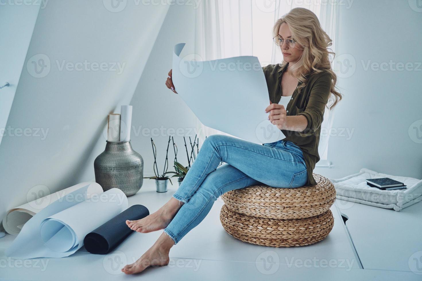Attractive young woman exmining blueprint while sitting on padded stool at home photo