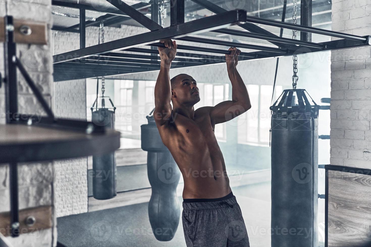 Challenging himself. Handsome young African man doing pull-ups while exercising in the gym photo