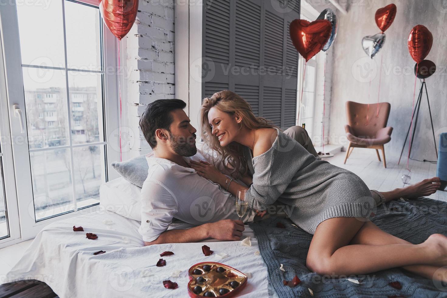 Sweet candies in bed.  Beautiful young couple eating chocolate candies and smiling while lying on the bed at home photo