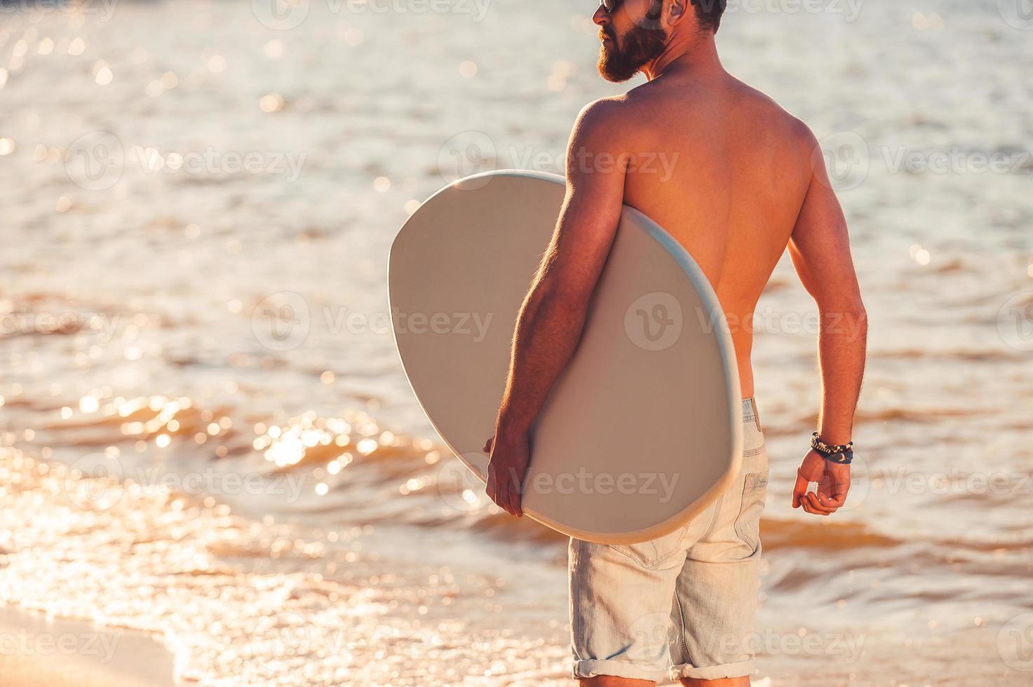 Lord of wave. Cropped image of shirtless young man holding skimboard and looking away while walking along the beach photo