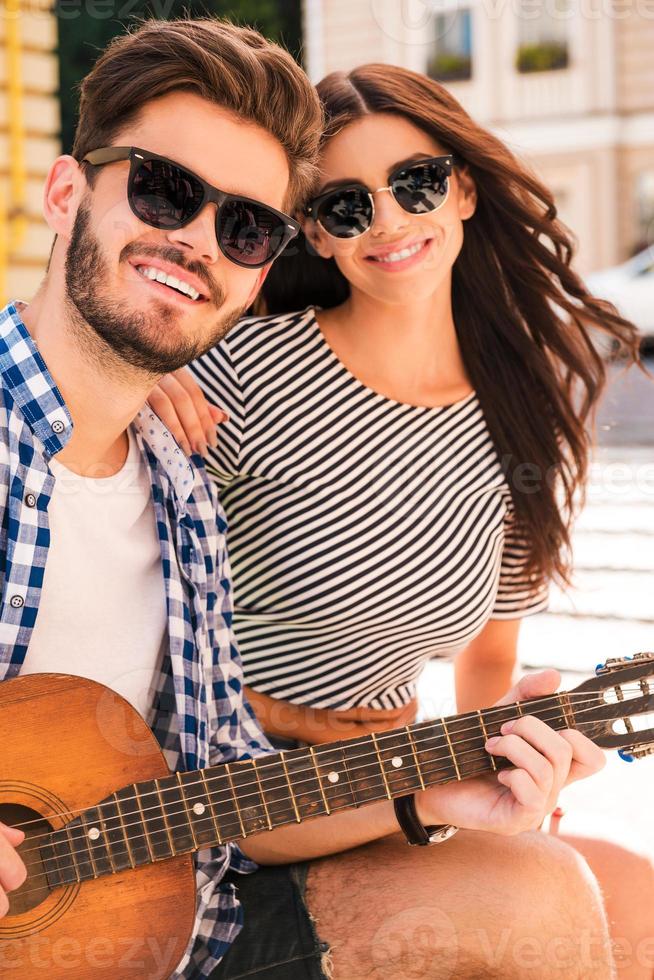 Spending carefree time together. Smiling young loving couple bonding to each other and looking at camera while sitting outdoors with guitar photo