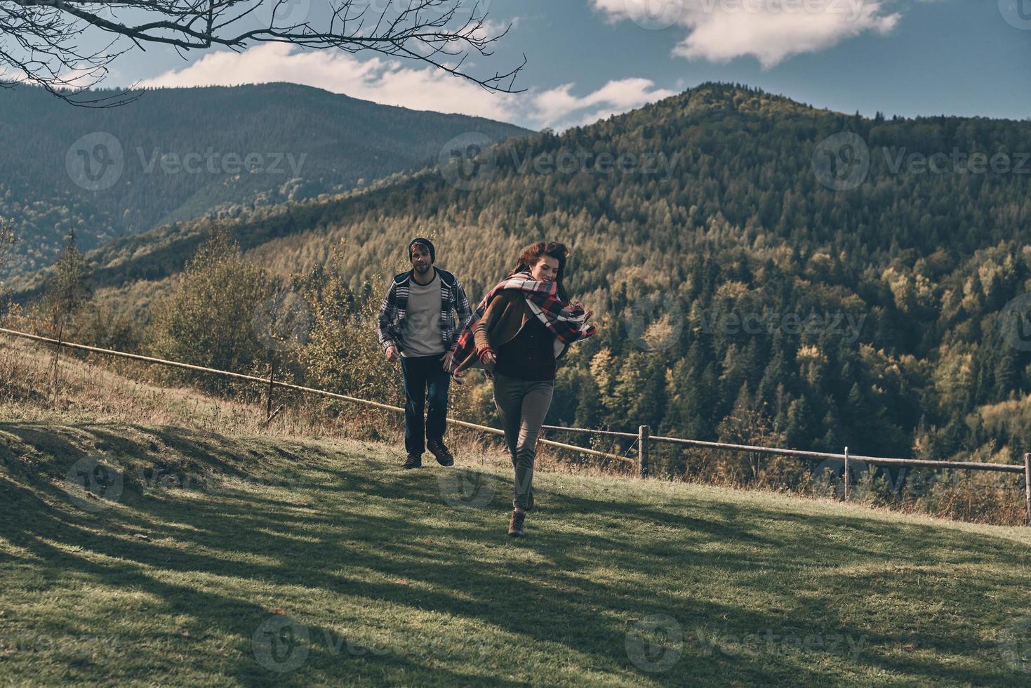 se siente como volar. Longitud total de feliz pareja joven sonriendo mientras corre en el valle en las montañas al aire libre foto