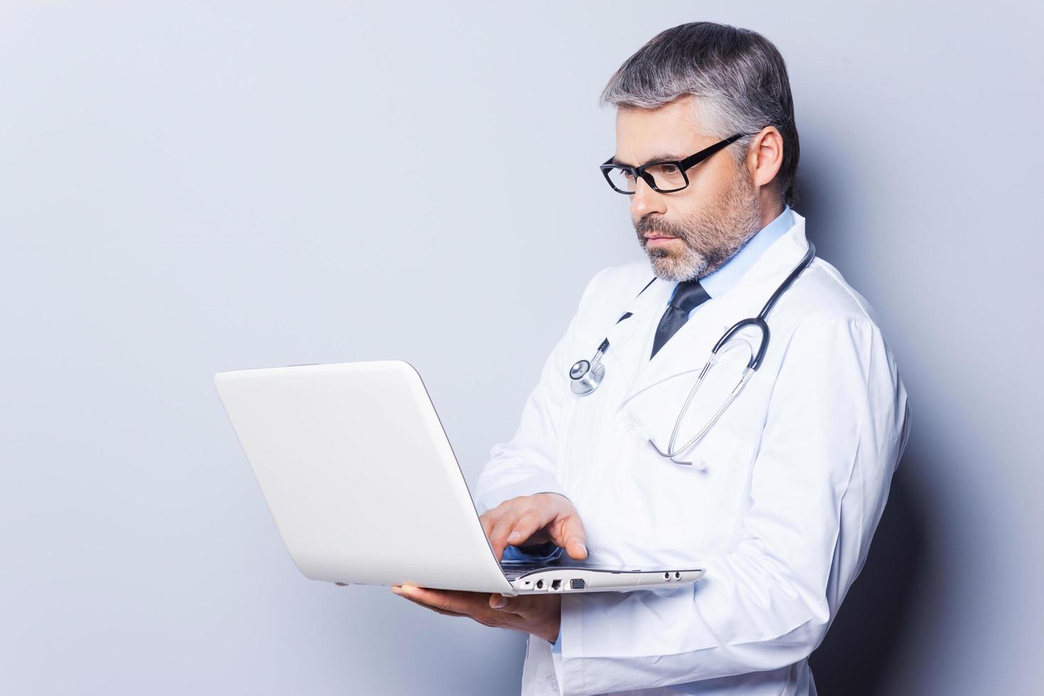 Doctor with laptop. Confident mature doctor working on laptop while standing against grey background photo