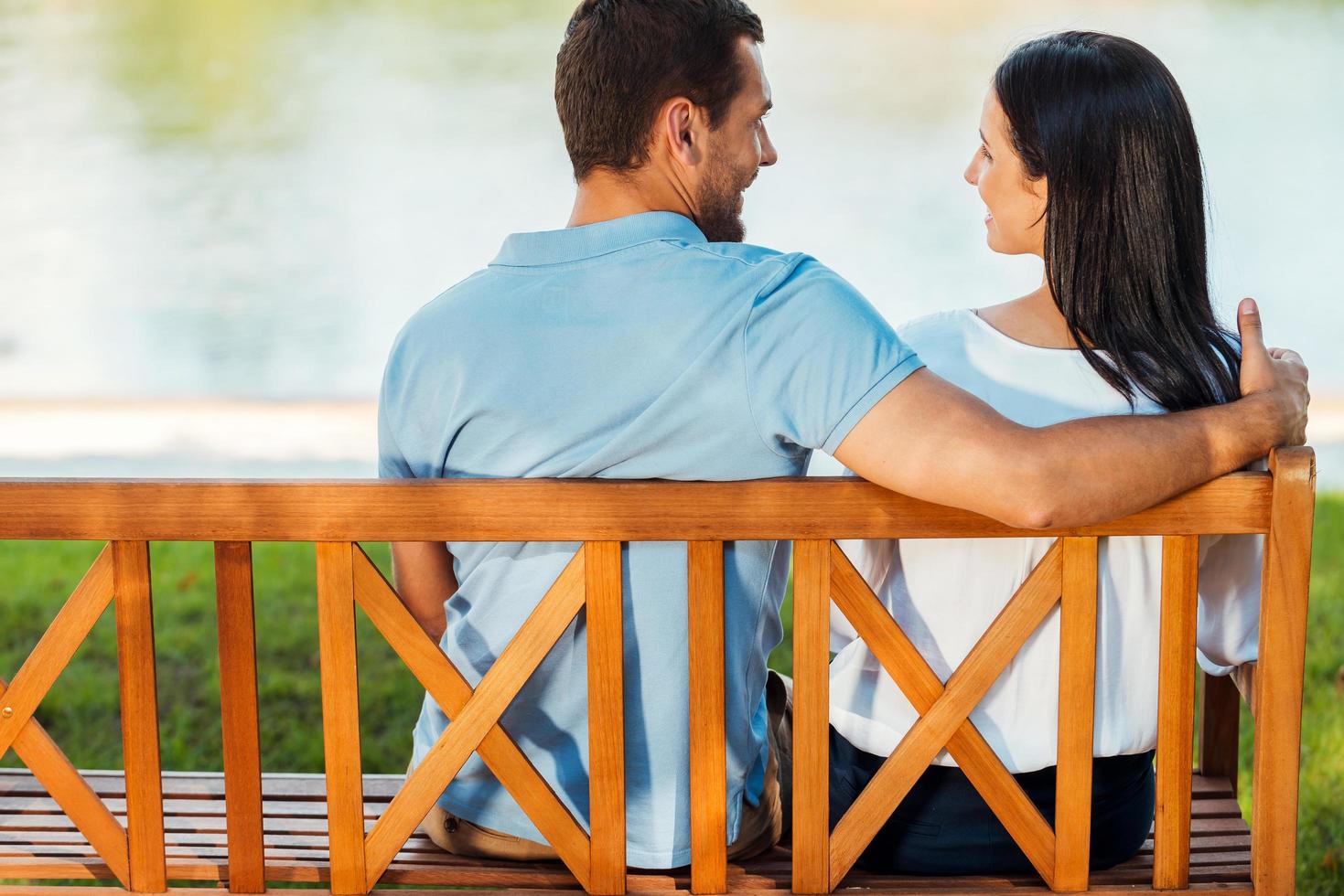 cita romántica. vista trasera de una hermosa joven pareja amorosa sentada en el banco juntos y sonriendo el uno al otro foto