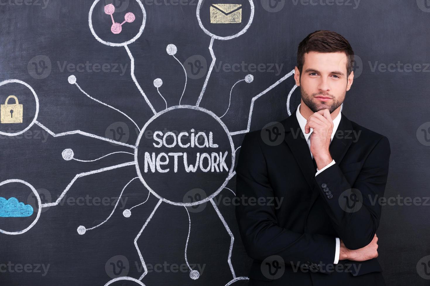 ser una persona activa socialmente. Apuesto joven sosteniendo la mano en la barbilla y mirando a la cámara mientras está de pie contra el dibujo de tiza de la red social en la pizarra foto